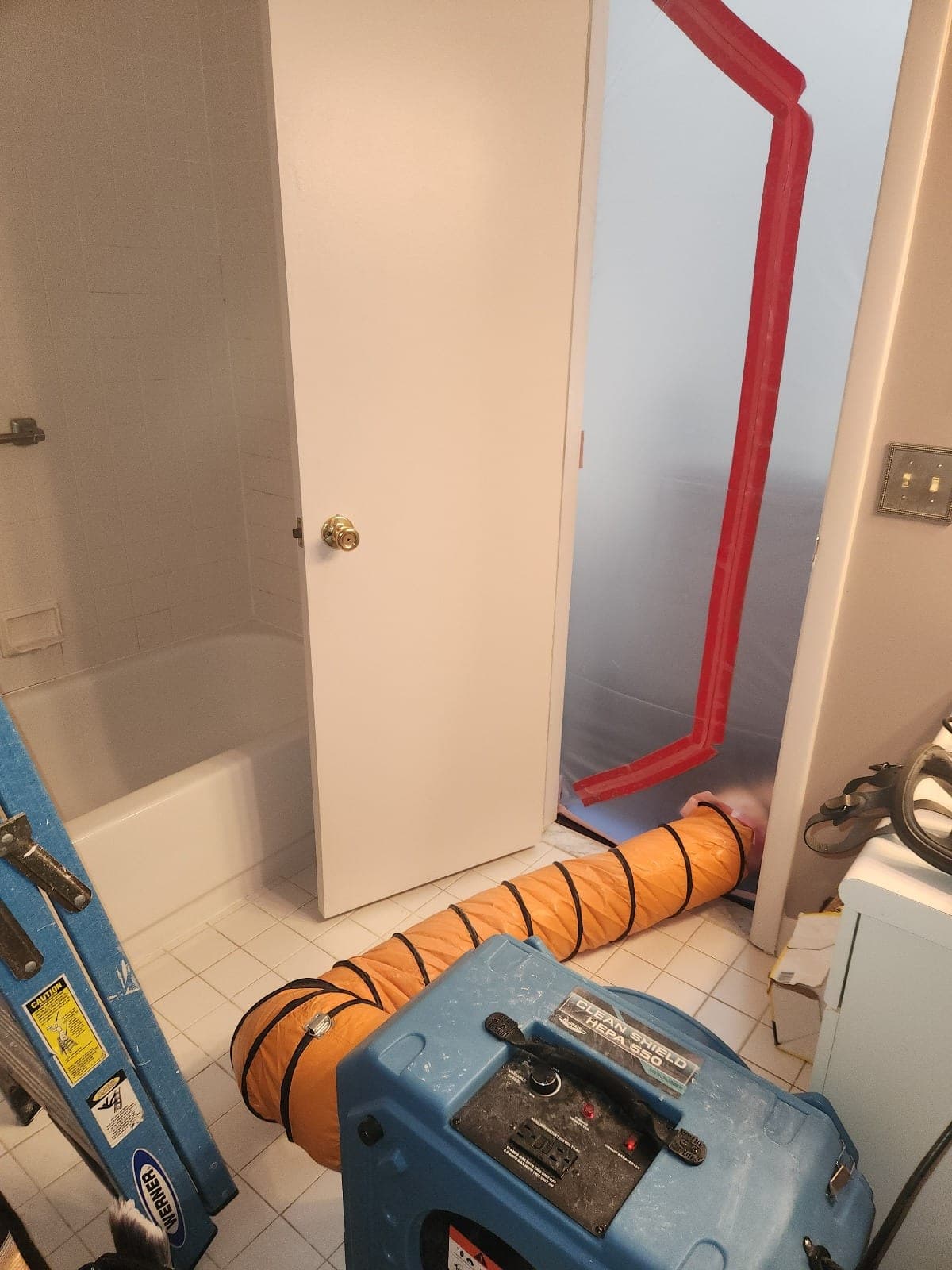 HEPA negative air machine with orange flex duct running into a poly-sealed doorway with red zip tape