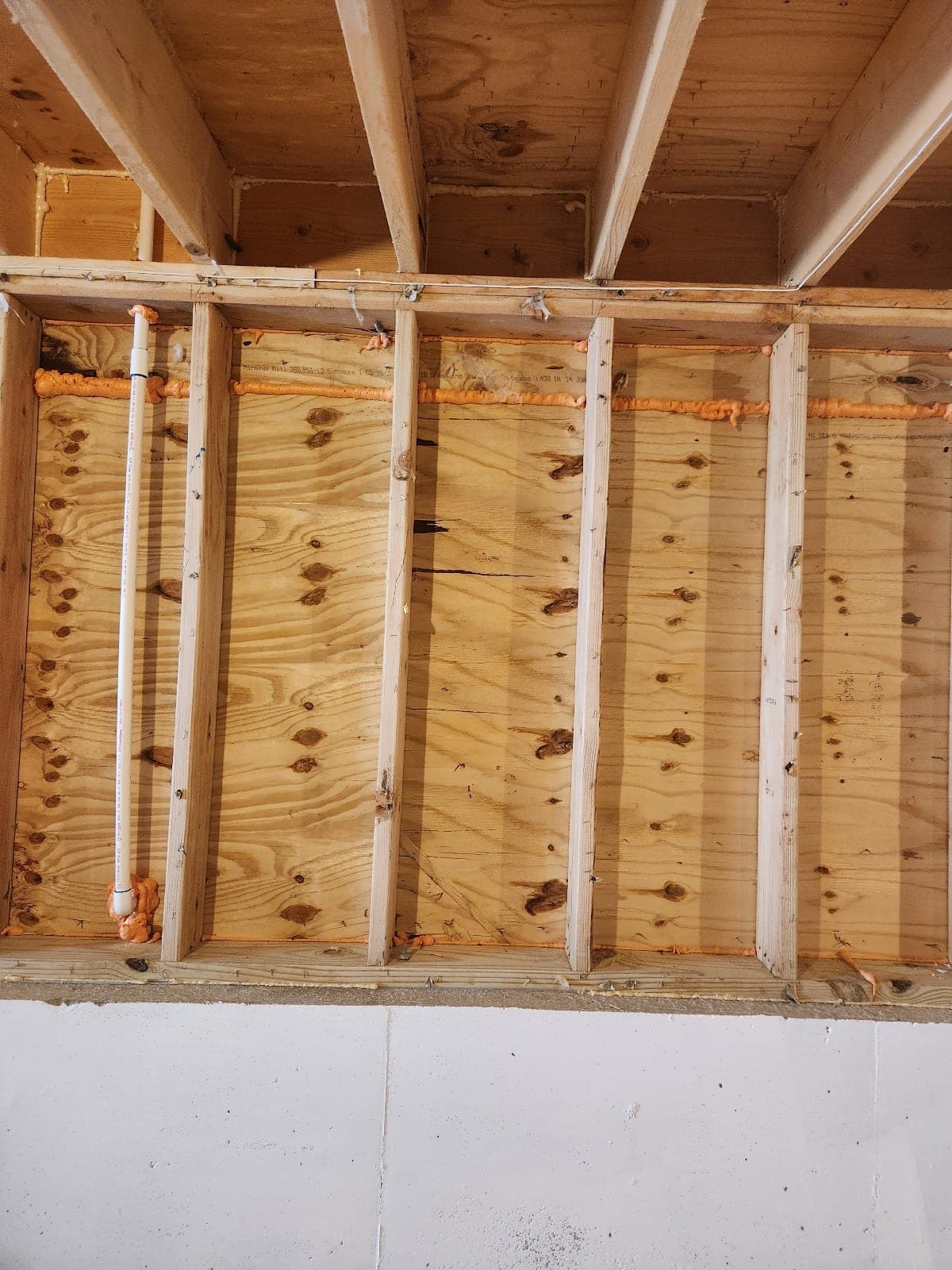 Open wall cavity with plywood sheathing, studs and fresh orange spray-foam sealant lines along the plates