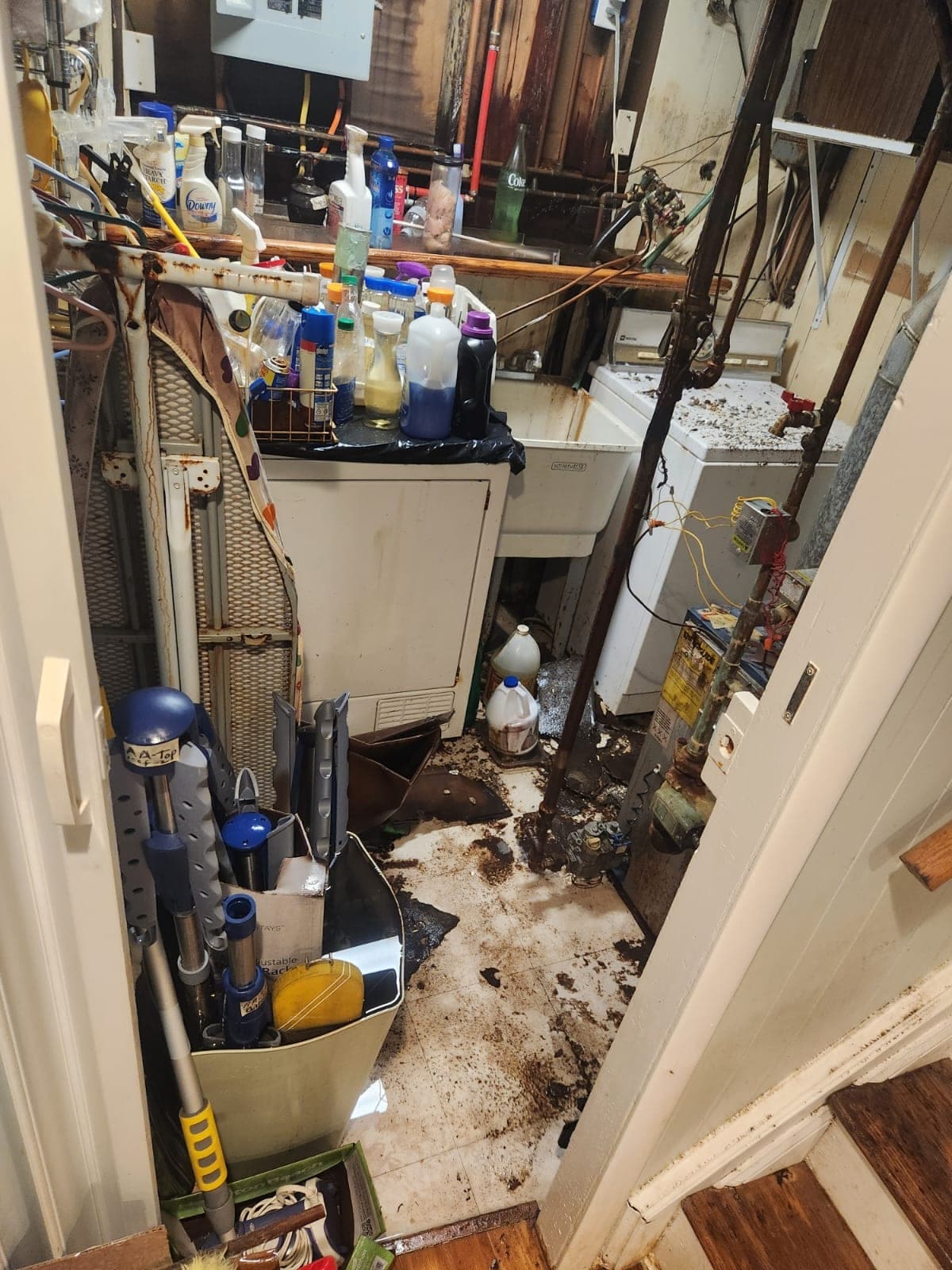 Cramped laundry room with dark sewage sludge spattered across the floor around a utility sink and washer
