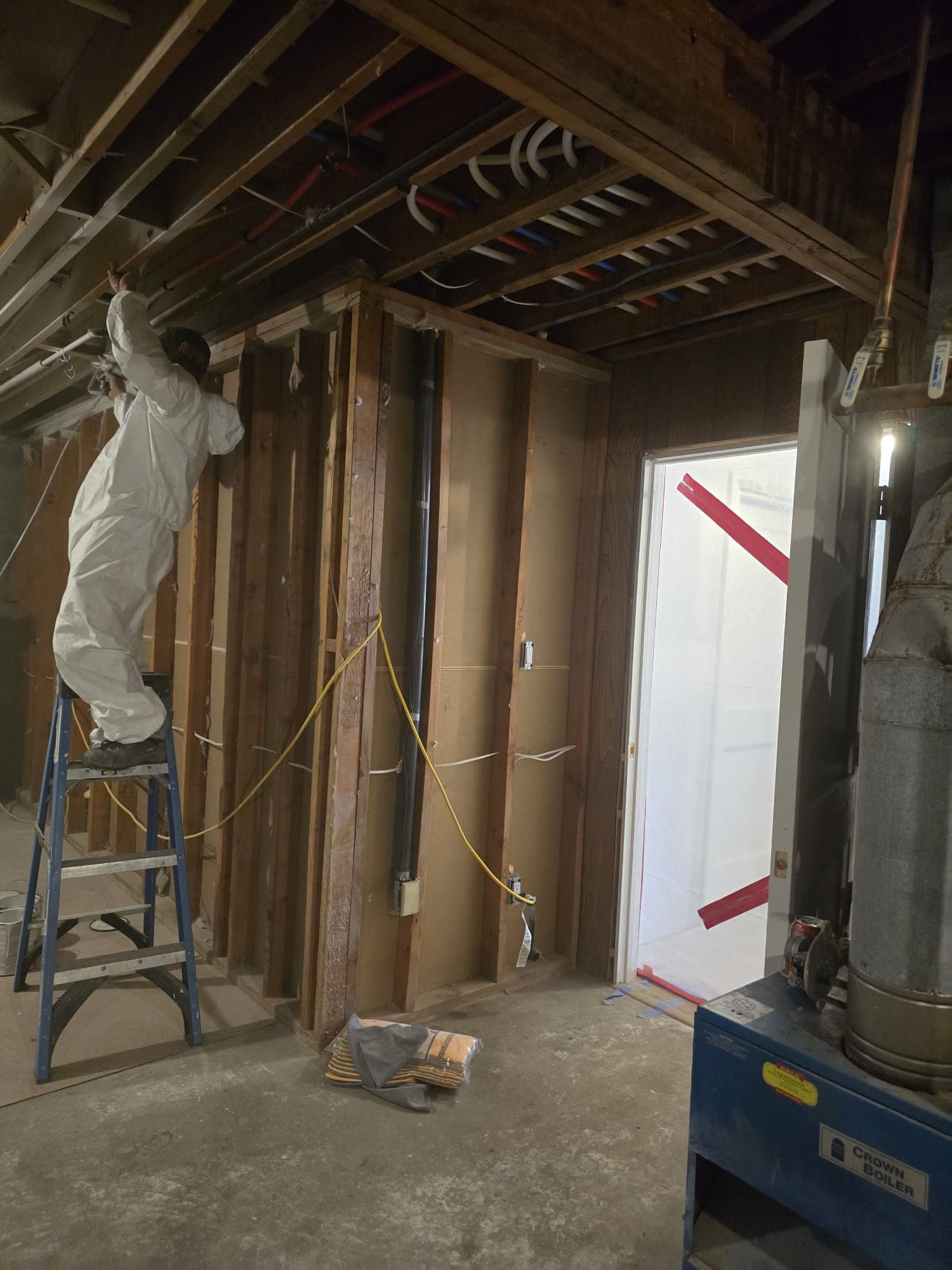Technician in white protective coveralls on a ladder treating exposed framing, red-taped containment door behind
